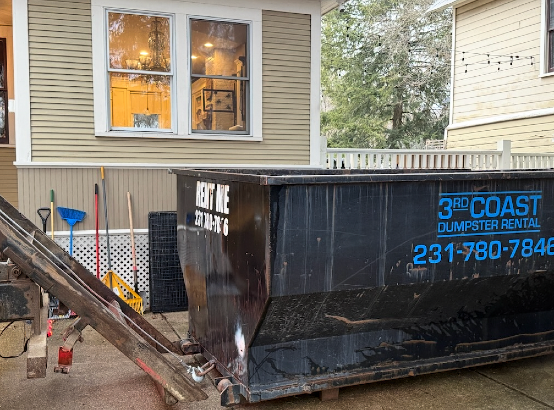 Large black dumpster beside a house, with a wooden ramp leading down to it and a window above.