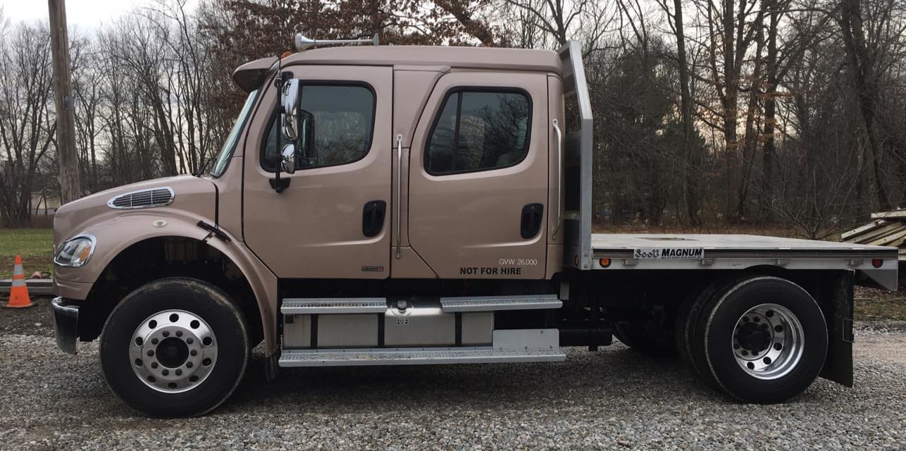 A truck with a flat bed is parked in a gravel lot.