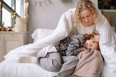 Woman cuddling a child under a white blanket on a bed in a cozy bedroom