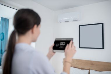 Person holding a tablet toward a wall-mounted air conditioner in a bright bedroom