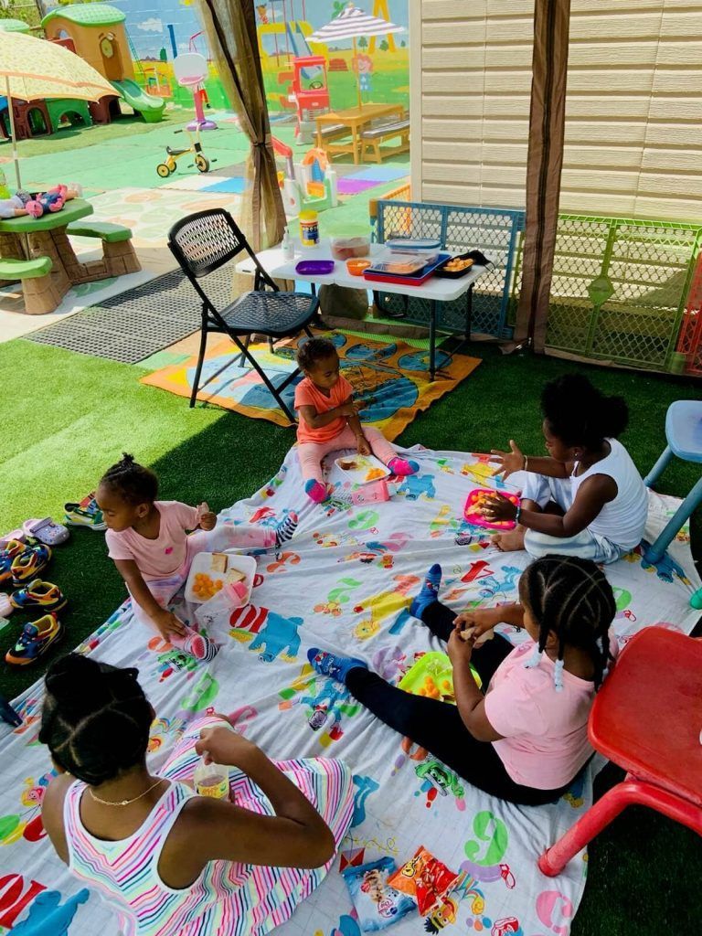 Children on a blanket playing outside, surrounded by toys and a colorful playground.