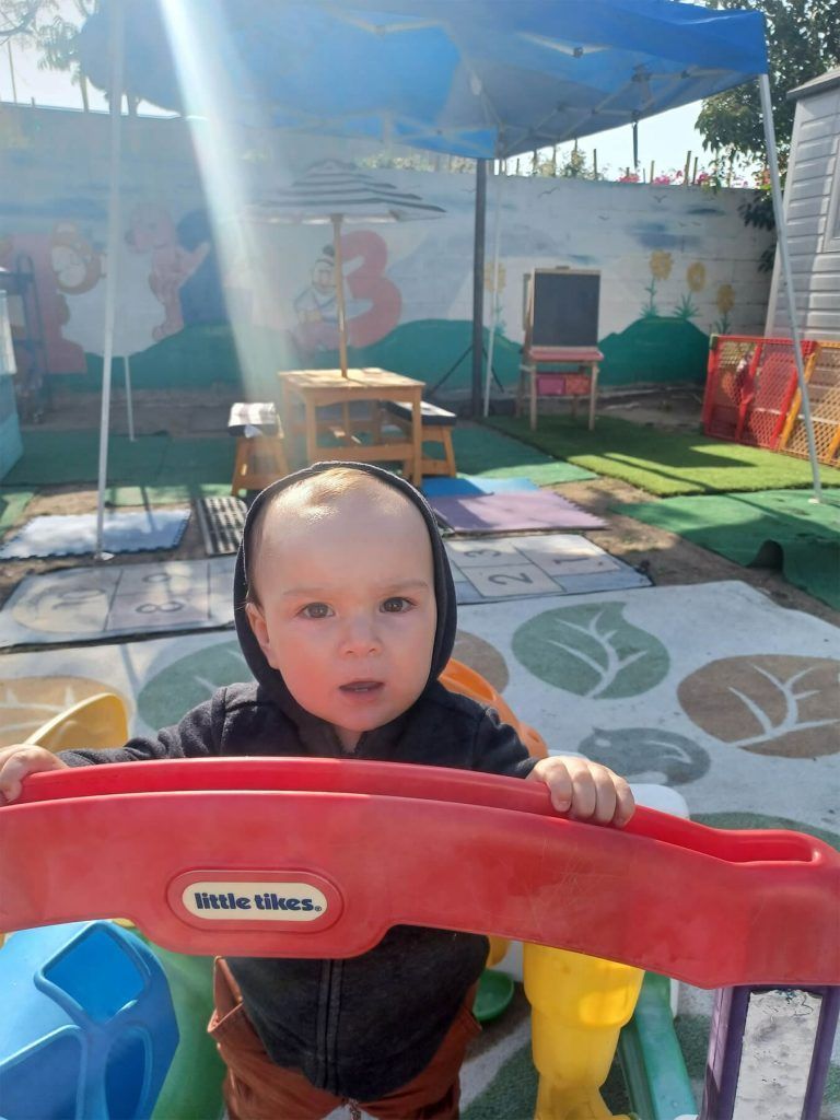 Child in a black hoodie holding a red toy, looking at the camera in a sunny outdoor play area.
