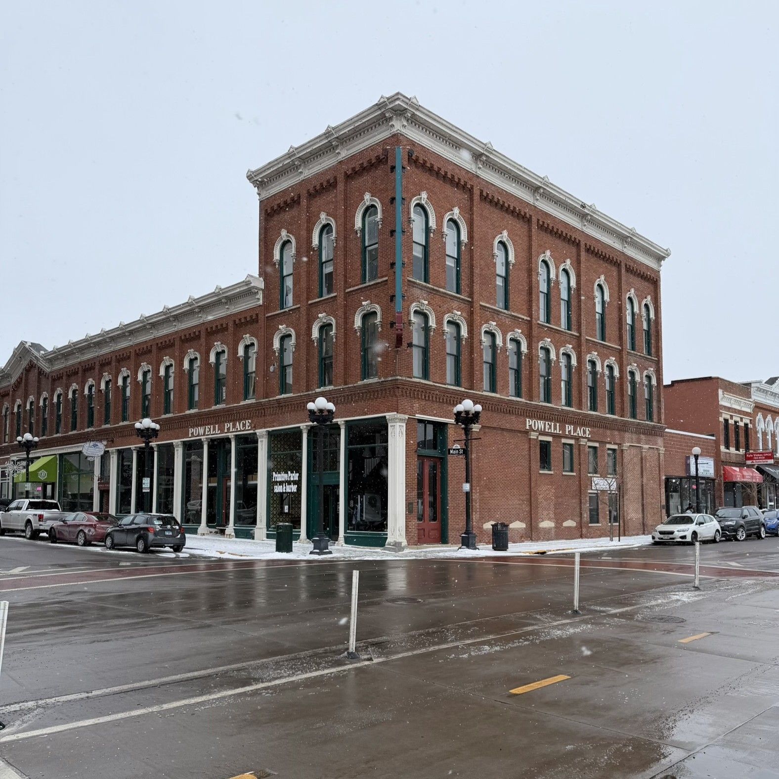 Snowy street corner with a red-brick historic building and storefronts on a wet road