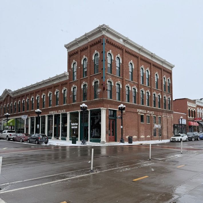Snowy street corner with a red-brick historic building and storefronts on a wet road