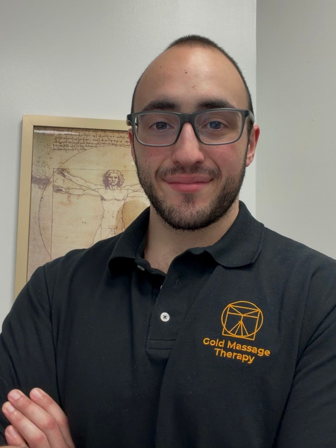 Person in glasses and black polo with Gold Massage Therapy logo, standing with arms crossed and smiling.