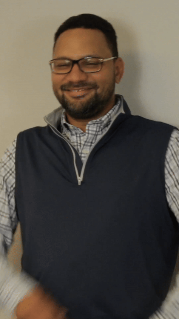 A man wearing glasses and a blue vest is smiling