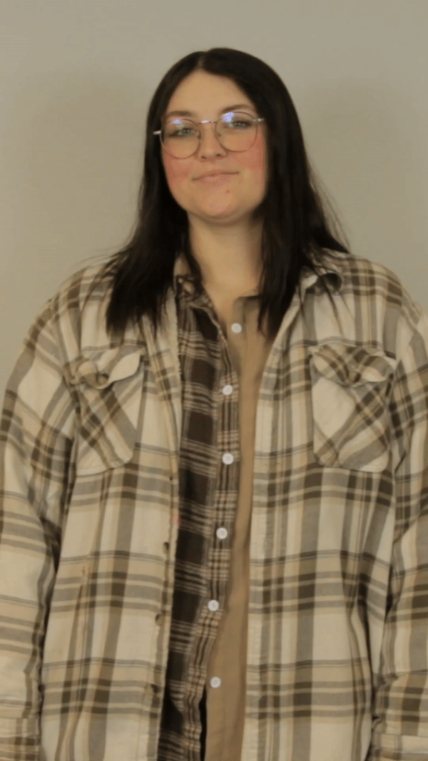 A woman wearing glasses and a striped sweater is standing in front of a white brick wall.