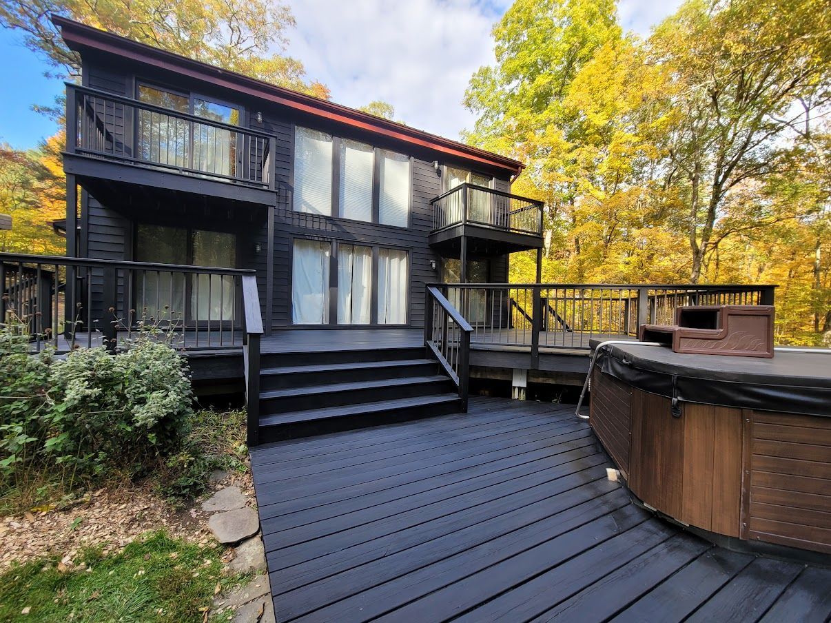 Black two-story house with deck, hot tub, and fall foliage in the background.