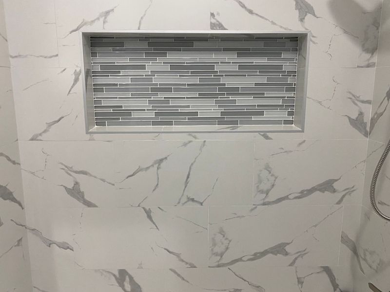 White marble tiled shower wall with rectangular recessed shelf tiled with gray mosaic.