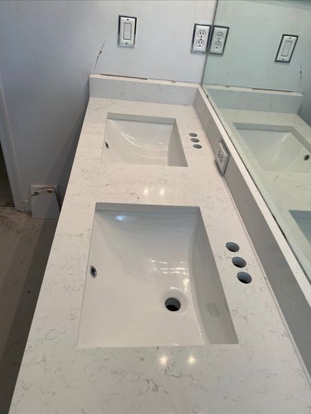 Double vanity with two white sinks, light countertop, and mirror. Wall-mounted light switches and outlets above.