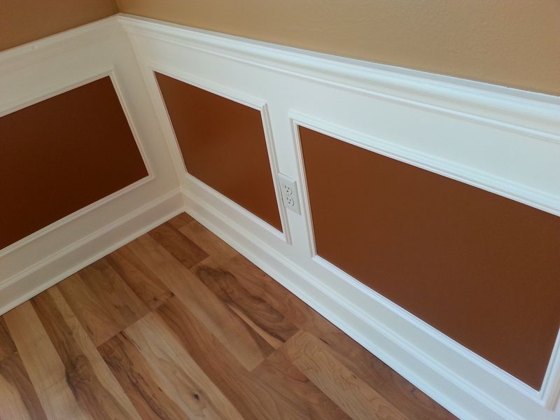 Corner of room with brown panel wainscoting framed in white molding, with a power outlet. Light wood flooring.