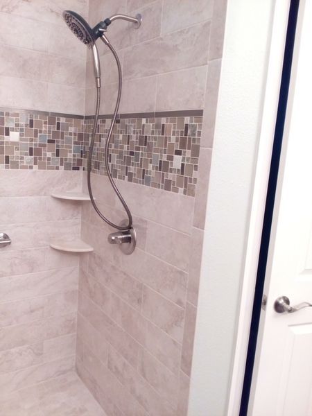 Shower with neutral tile and mosaic accent. Two shelves and a shower head. Doorway on the right.