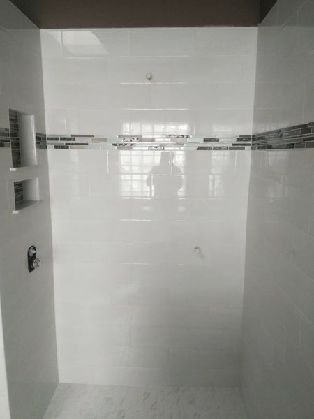 White-tiled shower with a decorative mosaic band and built-in shelf. A person is reflected in the shower's glass.