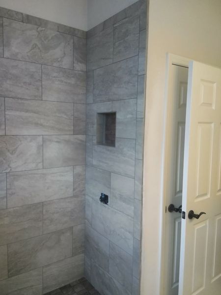 Corner of a tiled shower with a recessed shelf and an open door to a white hallway.