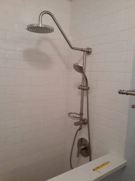 Shower with rain shower head, handheld shower, soap dish, and wall-mounted plumbing against white tiled wall.