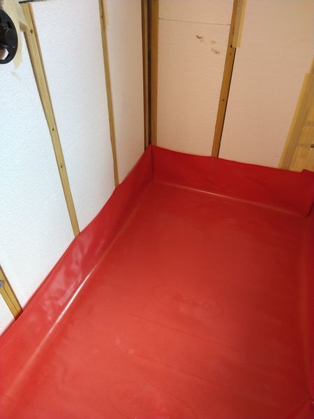 Red shower pan installed in a corner. Walls are lined with white panels and wooden studs are visible.