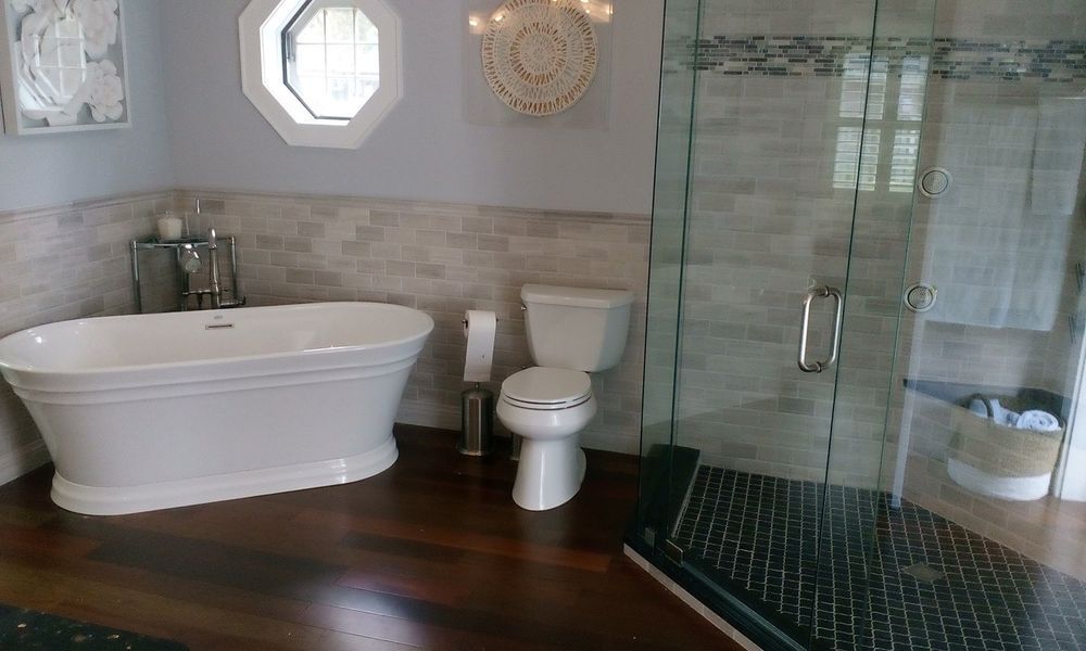 Bathroom with white soaking tub, toilet, and glass shower on wood floor.