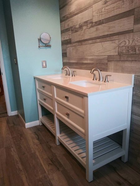 White double bathroom vanity with two sinks, drawers, and open shelves, against wood-look tile wall.