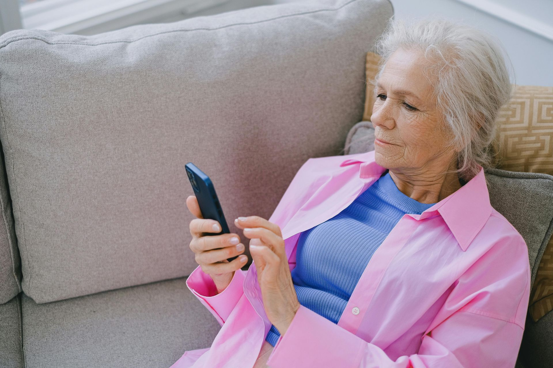 An elderly woman is sitting on a couch using a cell phone.