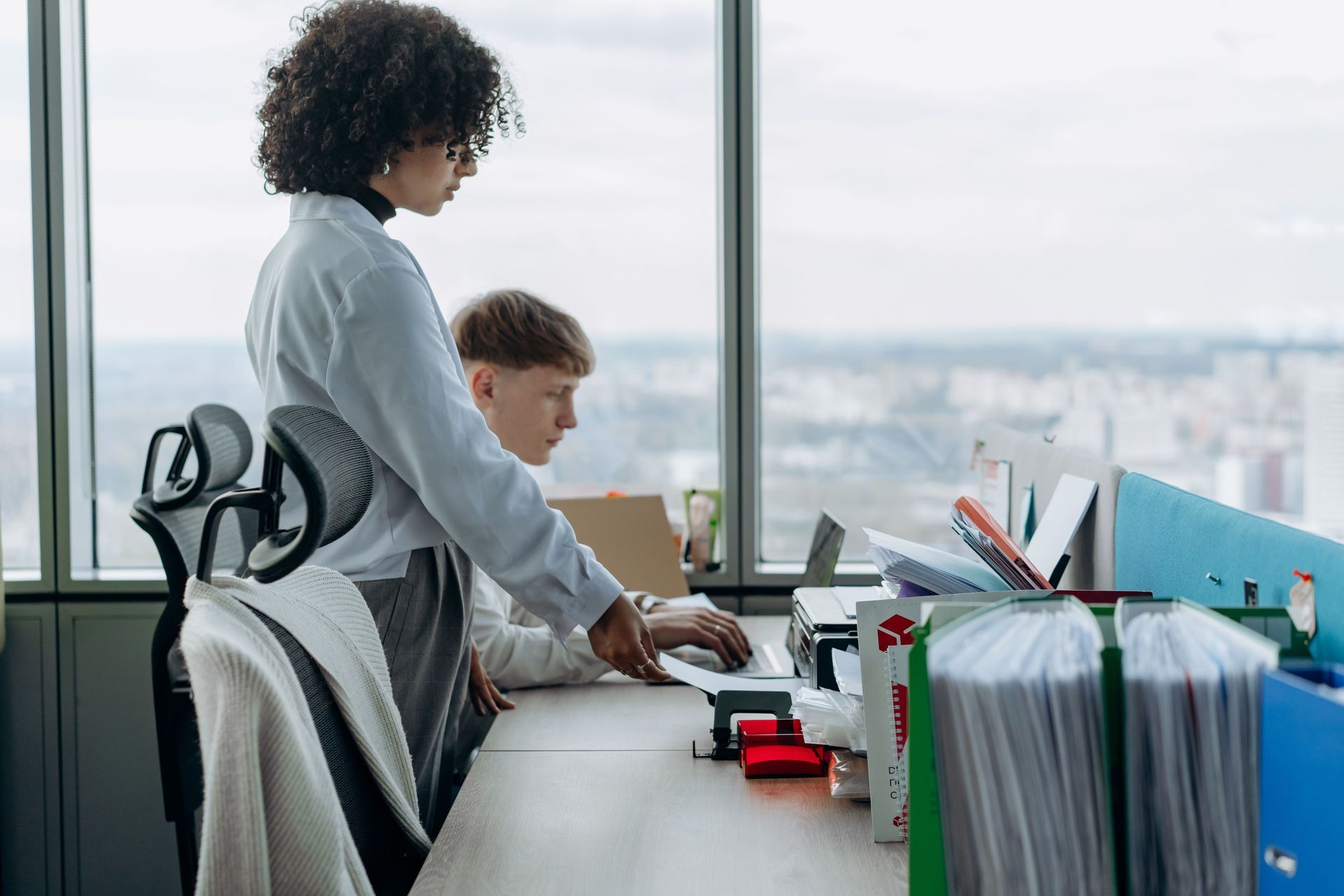 Two coworkers at a desk, one standing and typing while the other sits in front of a laptop in an office.