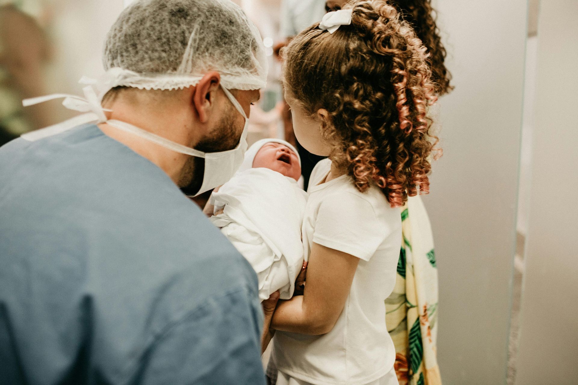 Doctor holding newborn, child watches, birthing room.