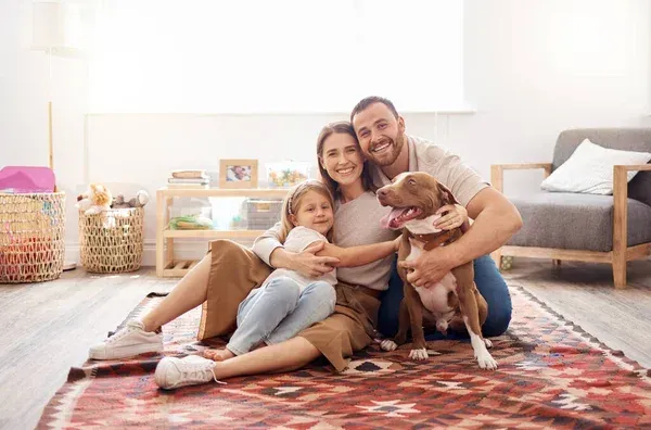 Family of three and dog sit on rug, smiling. Living room setting.