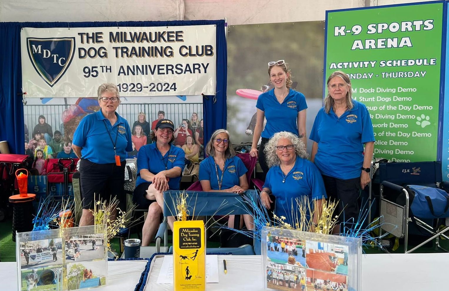 Milwaukee Dog Training Club booth with six women in blue shirts. Display includes a trophy, photos, and anniversary banner.