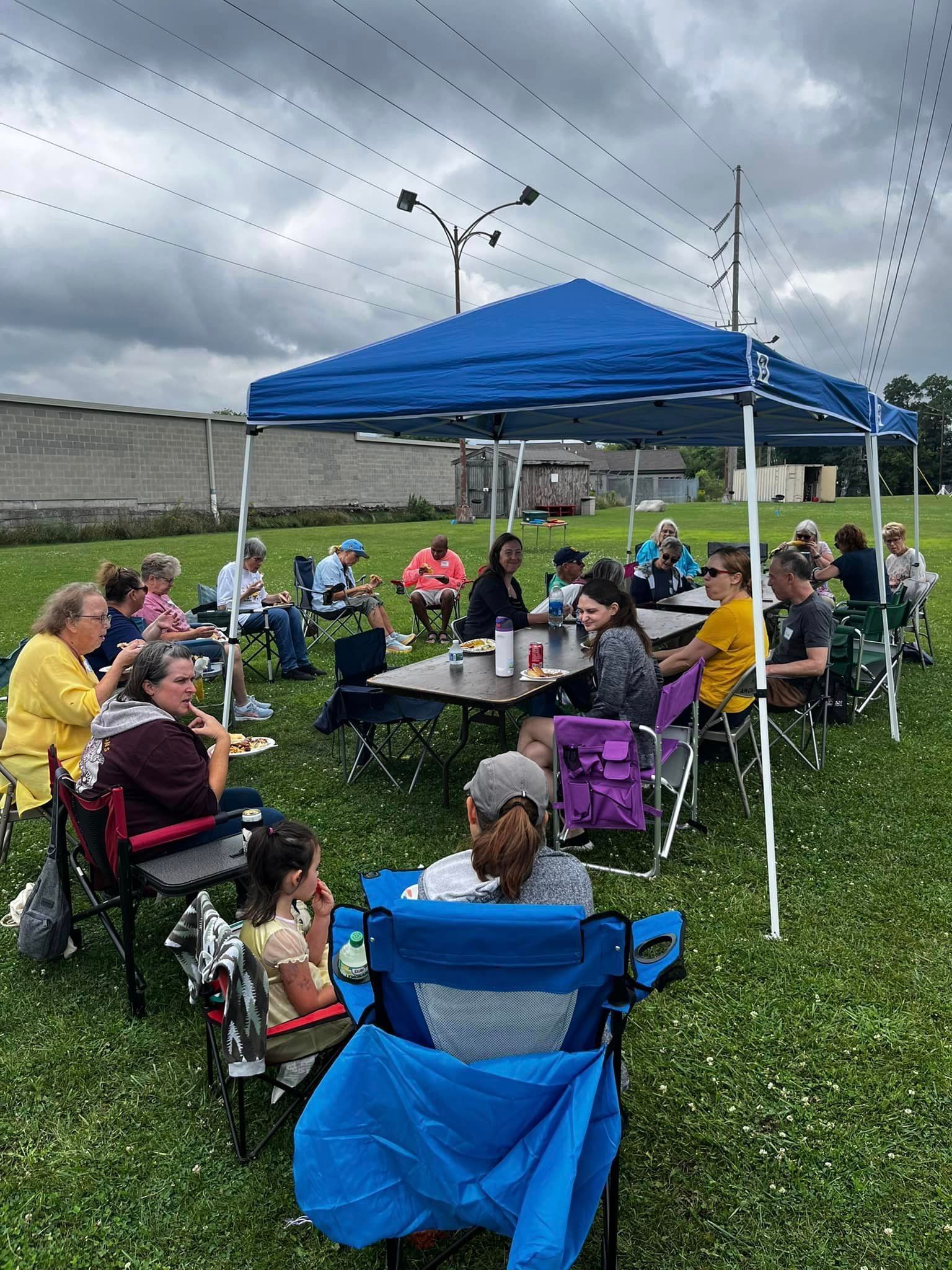 A group of people gathered under a blue canopy at an outdoor event. They are seated in folding chairs, eating and conversing on a cloudy day.