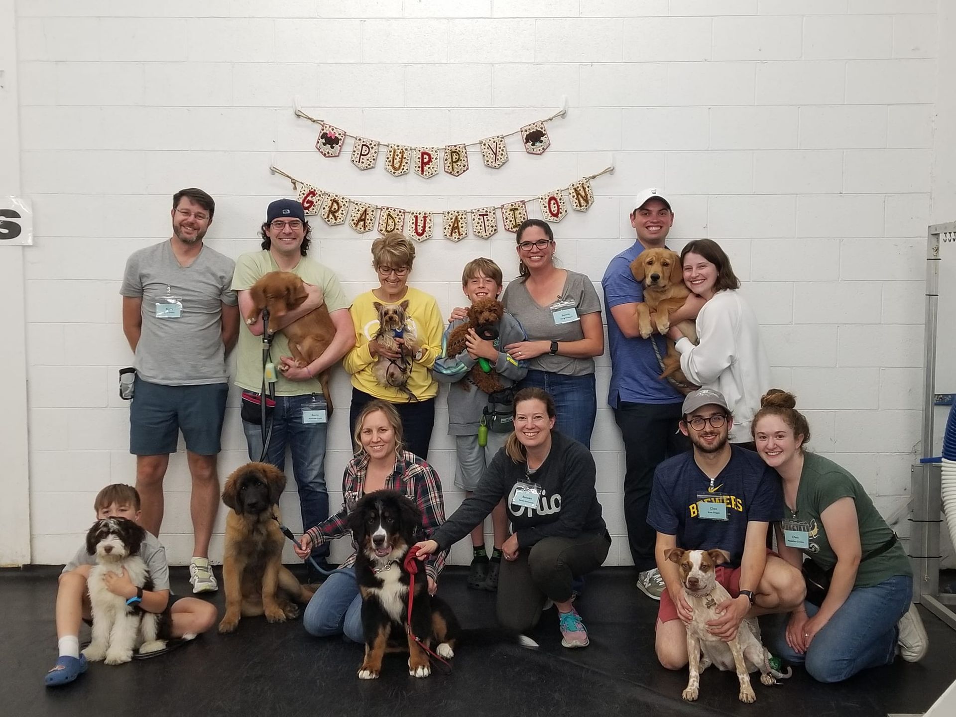 Group of people with dogs posing in front of a white wall decorated with a banner. Smiles and happy dogs.