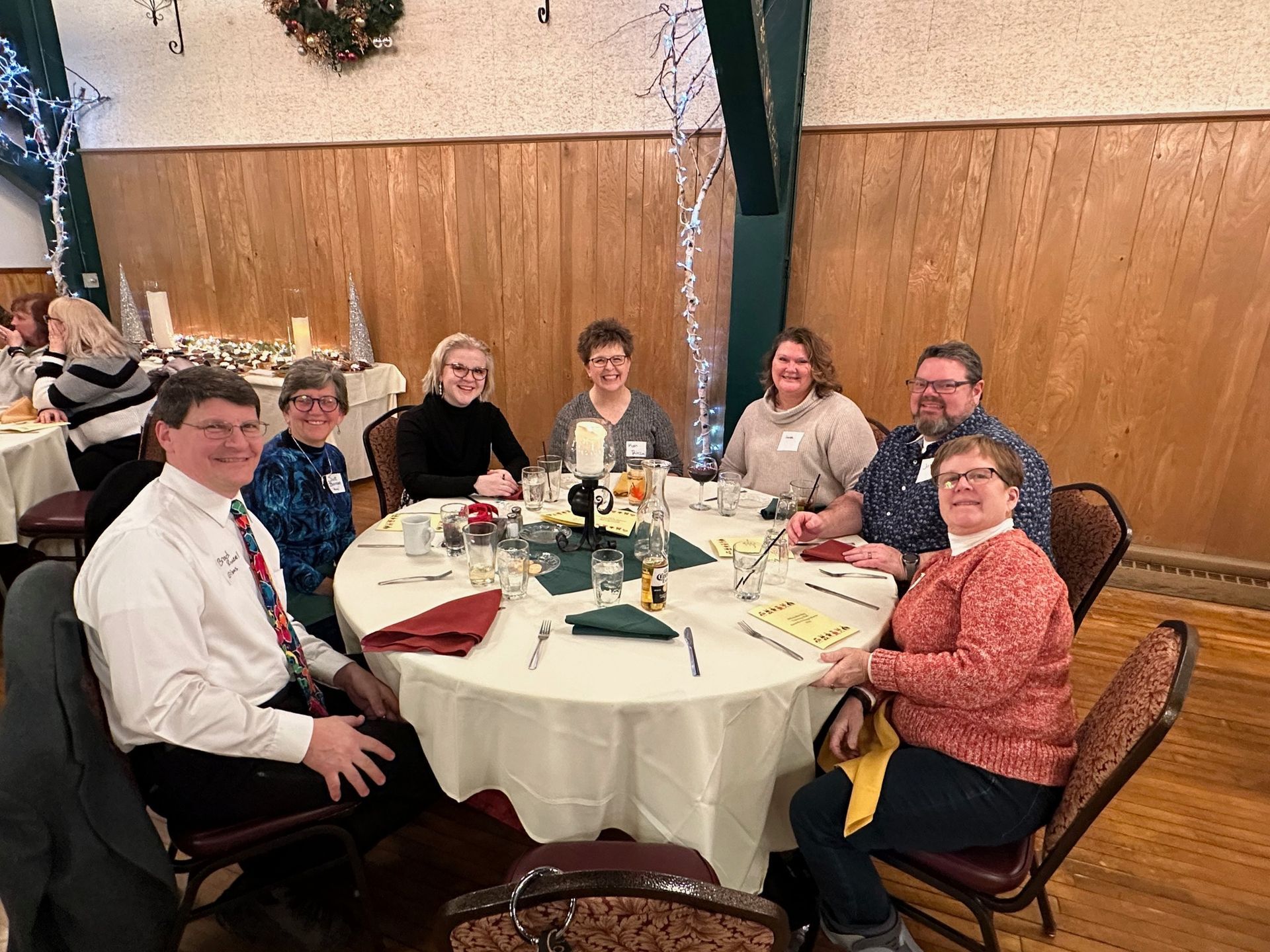 Group of seven people smiling around a decorated round table at a holiday gathering.