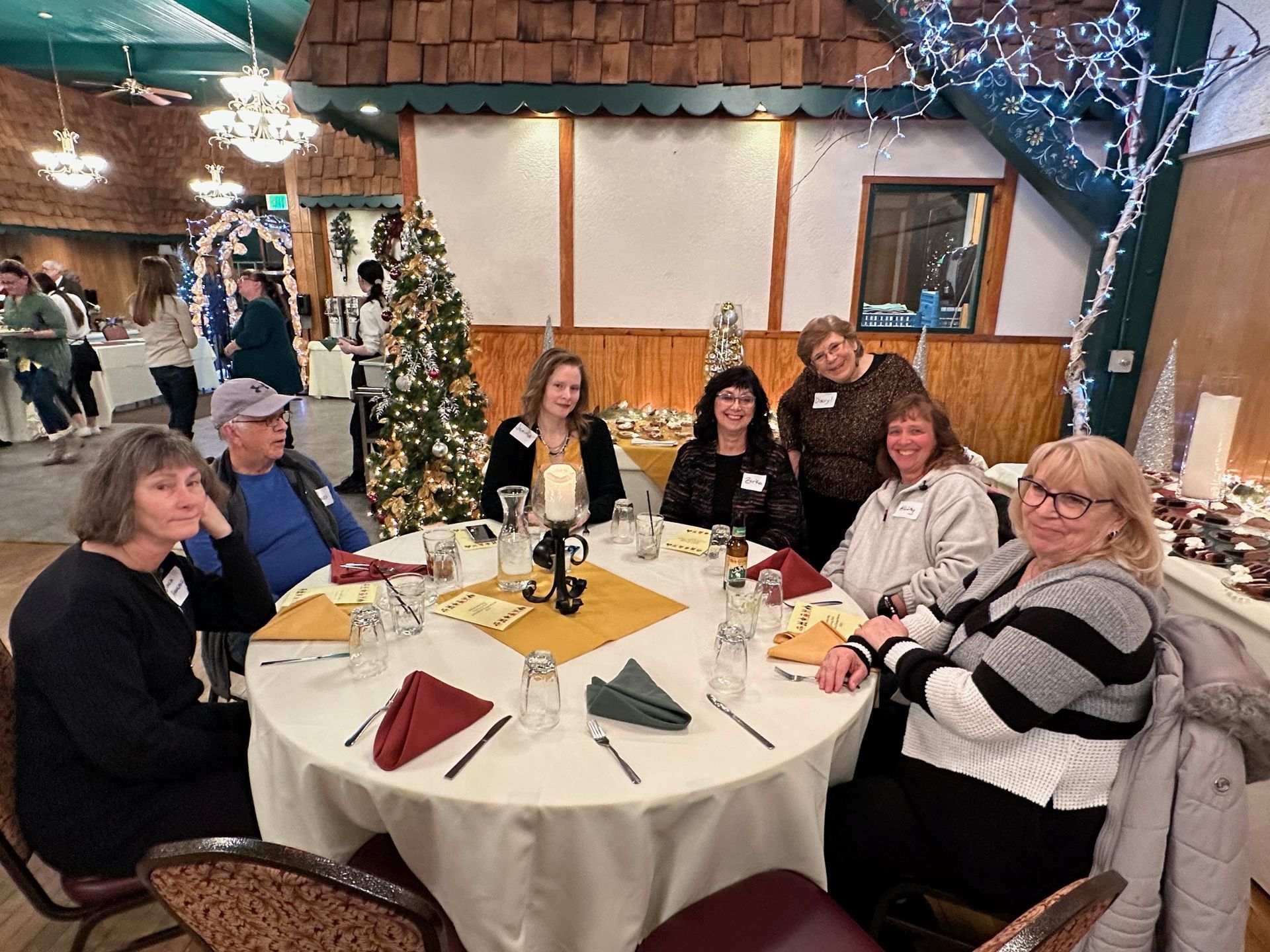 A group of seven people seated around a round table in a decorated room. They appear to be at a holiday gathering.