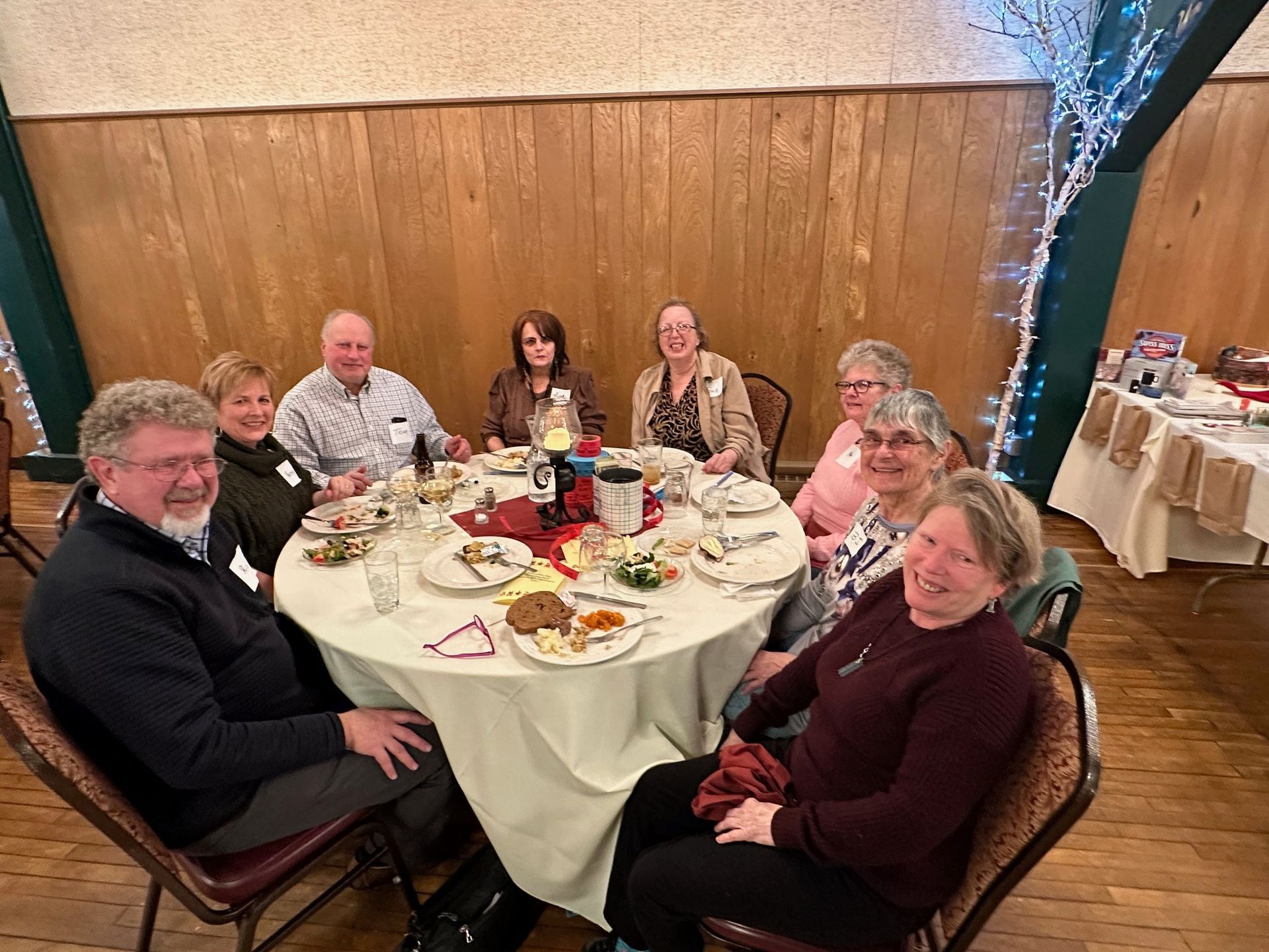 A group of nine people seated around a round table set for a meal, in a decorated room. People smile at the camera.