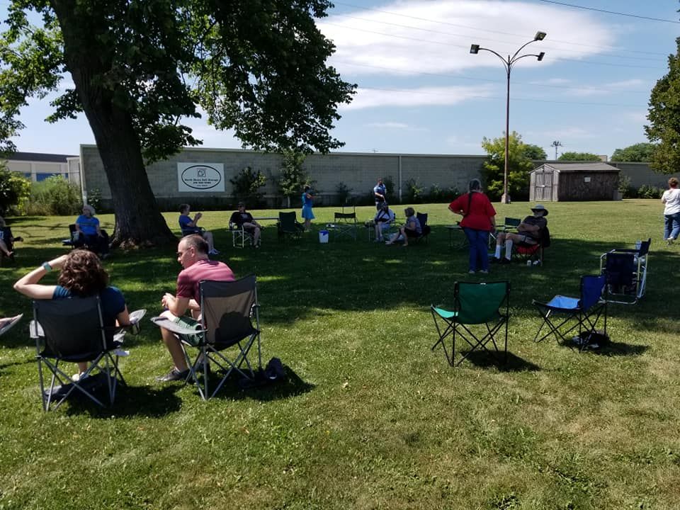 People sit in folding chairs on a grassy area under a tree, possibly an outdoor gathering or event.