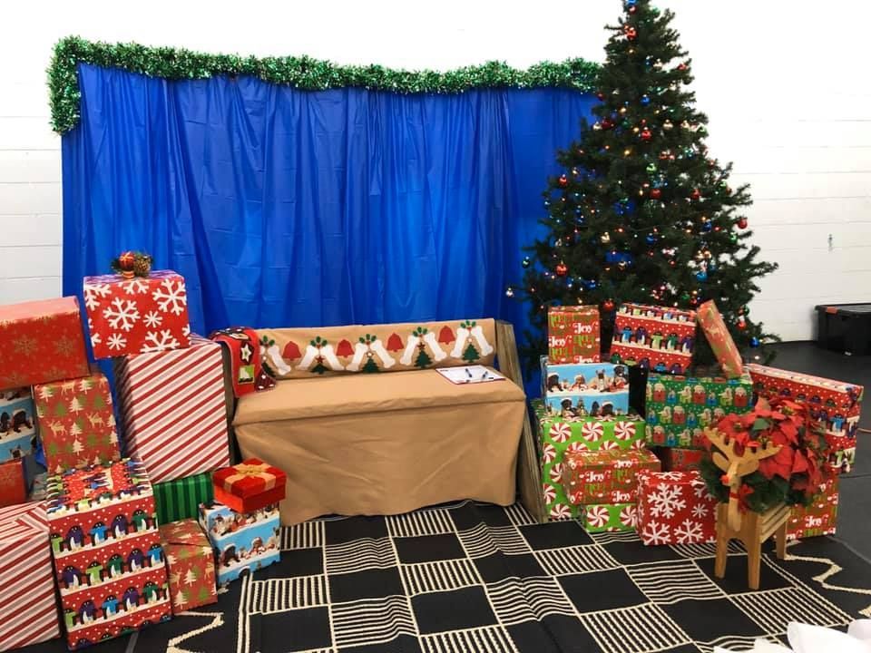 Christmas scene: Blue backdrop, decorated tree, wrapped gifts, a bench with a festive pillow, and a wooden reindeer.