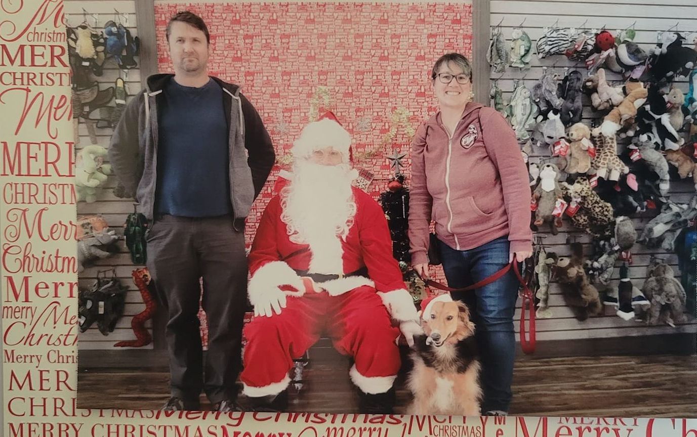 A couple and their dog pose with Santa Claus indoors. They are standing in front of a Christmas-themed backdrop.