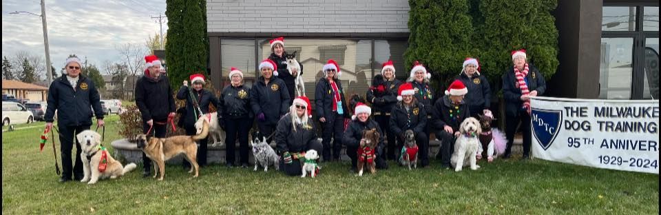 A group of people and dogs wearing Santa hats pose for a photo outside a building with a sign for dog training.