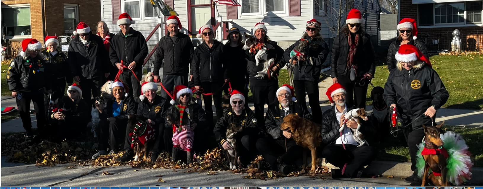 A group of people and dogs wearing Santa hats pose outdoors in front of houses; many are wearing black jackets.