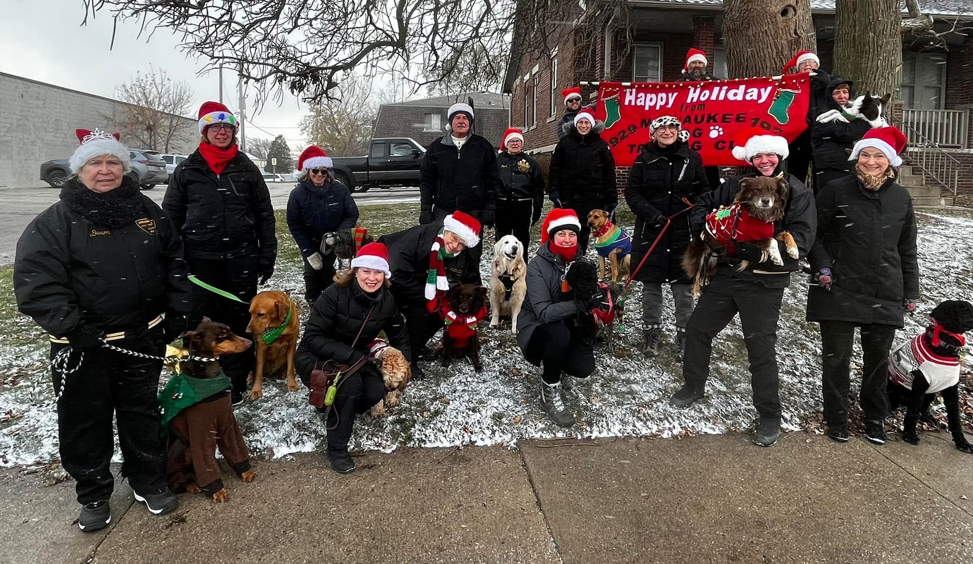 A group of people and dogs wearing Santa hats in a snowy outdoor setting. They are holding a 