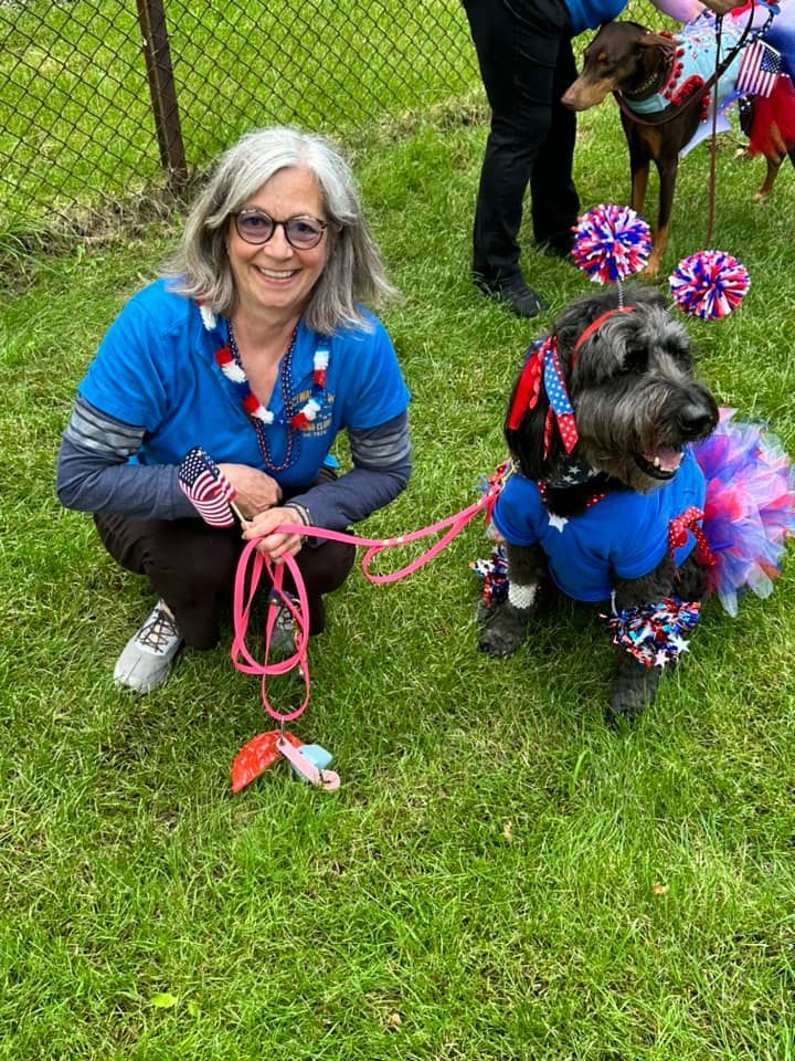 Woman and dog, both dressed in patriotic outfits, pose outdoors. The dog wears a tutu, and the woman smiles.