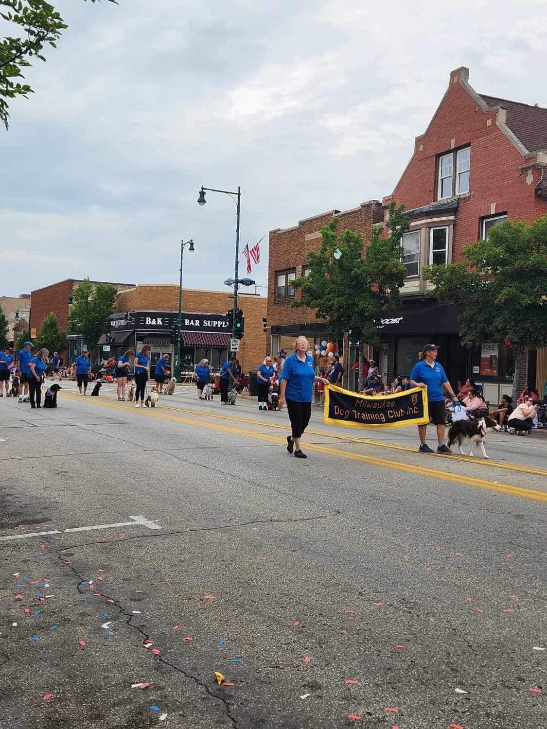 People in blue shirts walk with dogs in a parade on a city street. Buildings line the street, under a cloudy sky.