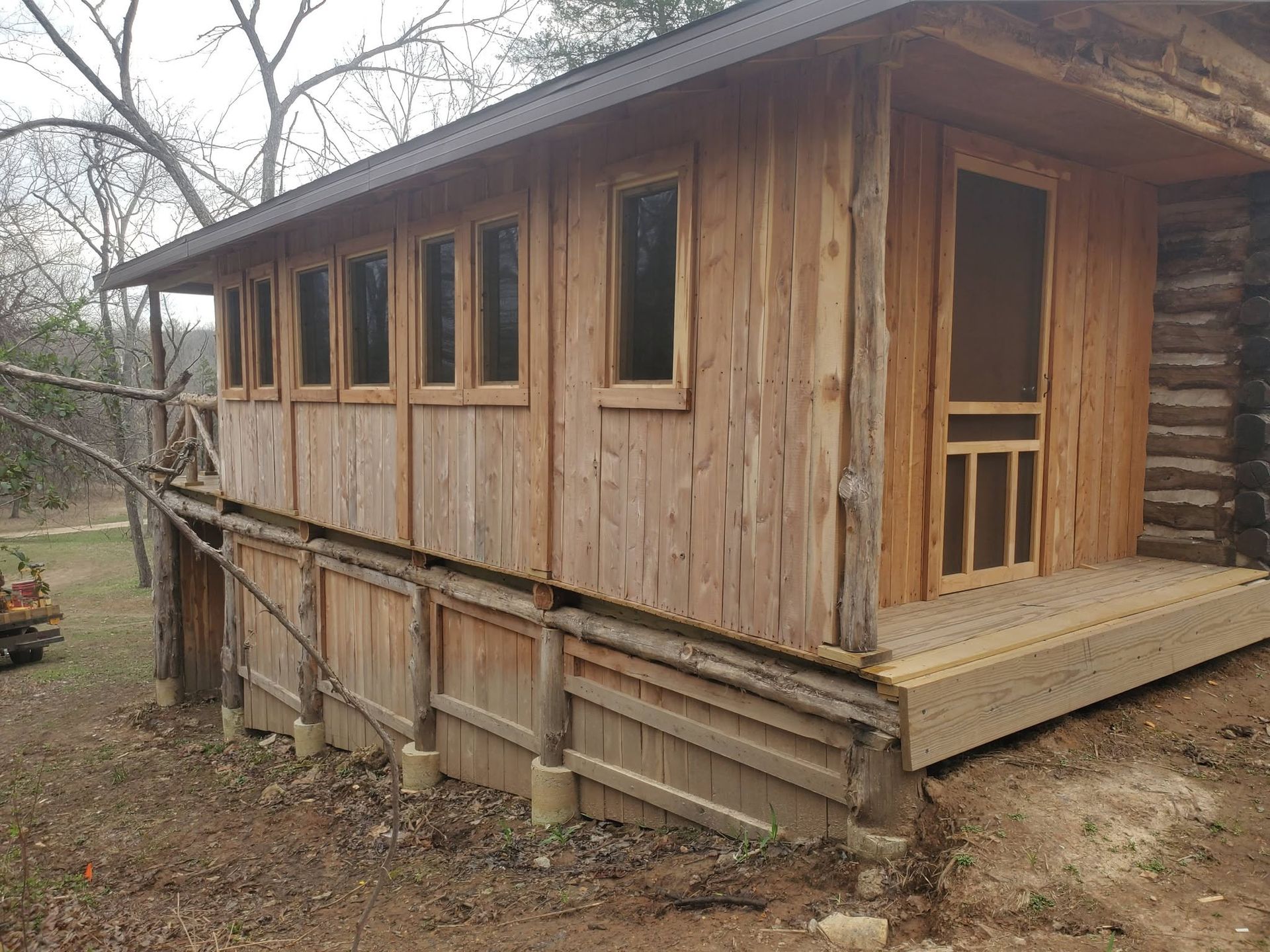 Wooden cabin with porch and ramp, viewed from the side in a wooded rural setting