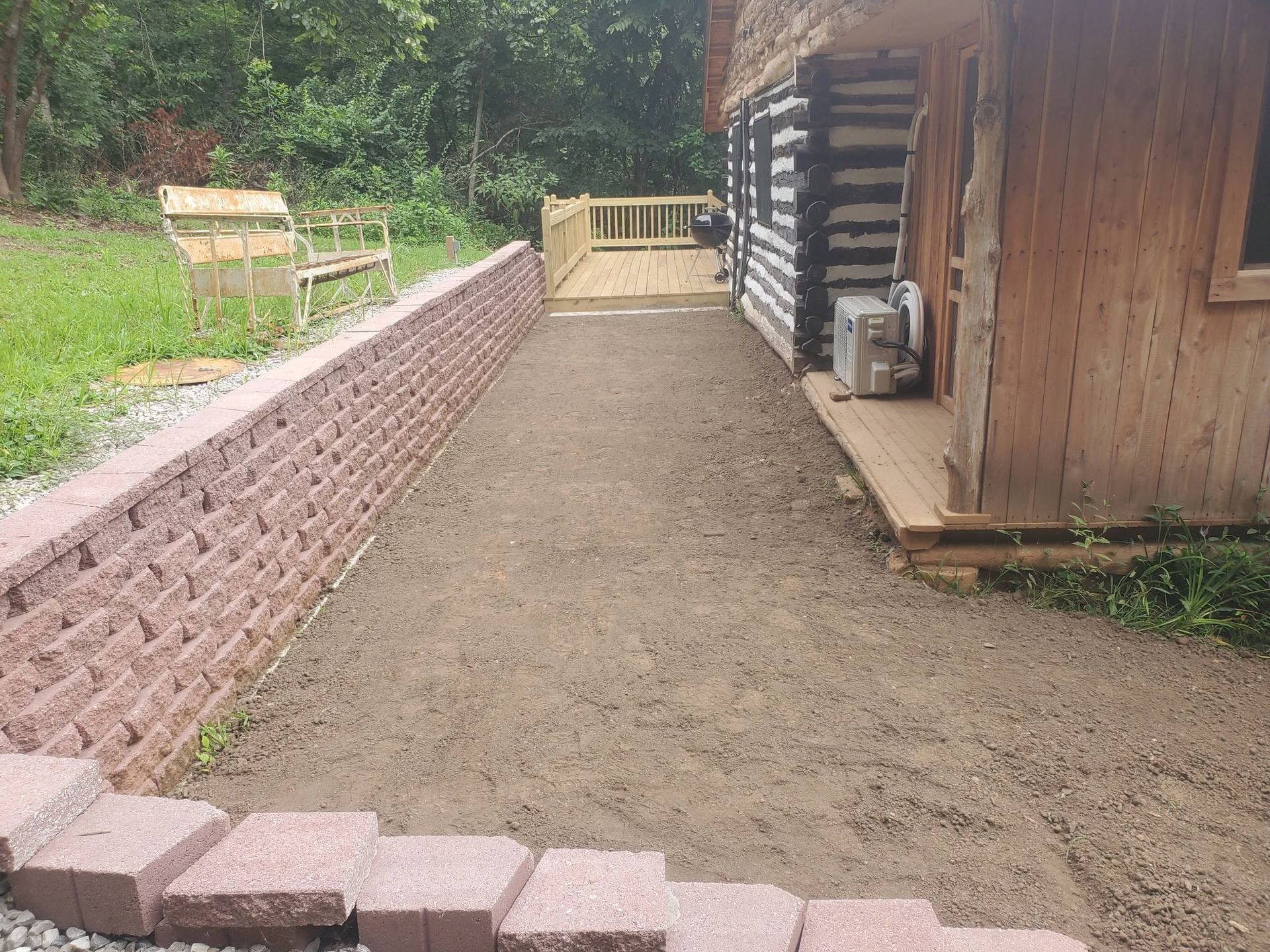 Gravel walkway beside a log cabin with a brick retaining wall and wooden bench in a wooded yard