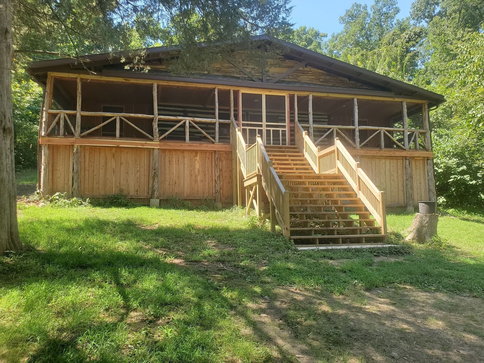 Wooden cabin with a front porch and central staircase on a grassy hillside under trees