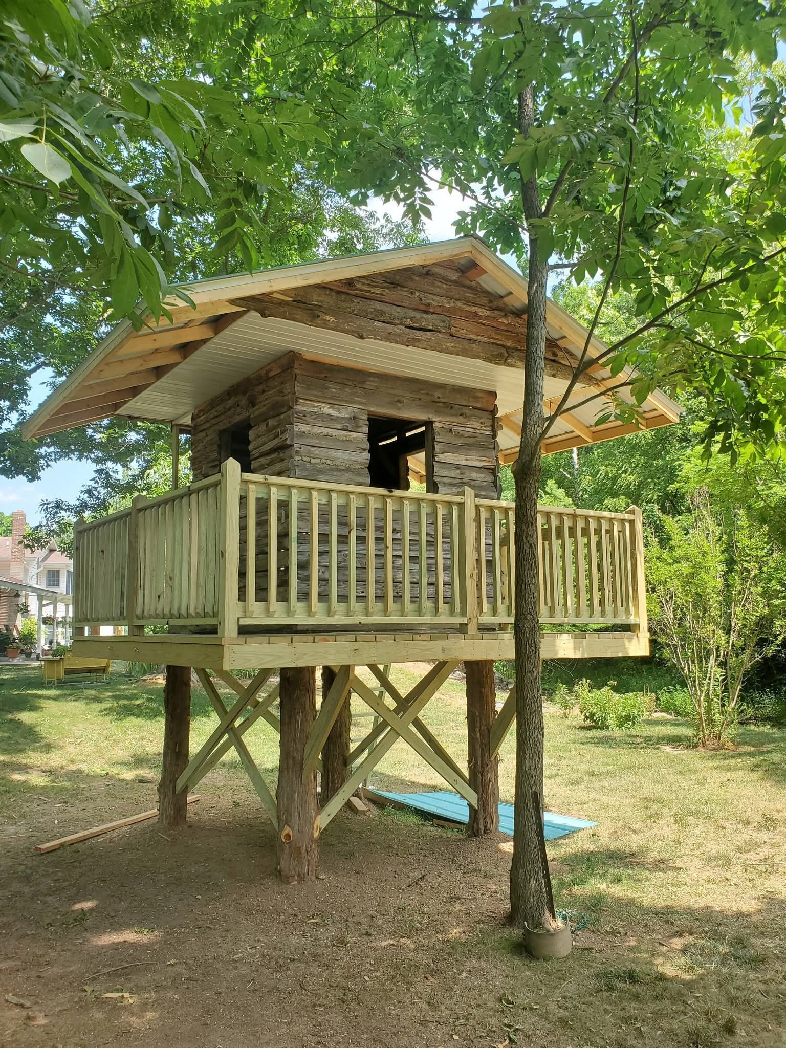 Small wooden treehouse on stilts with a wraparound deck in a leafy yard