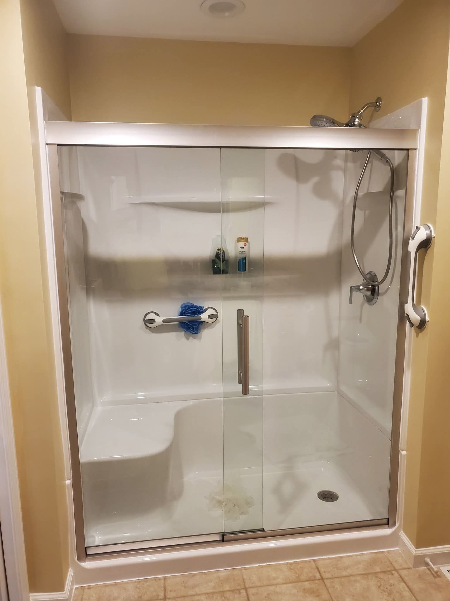 Walk-in shower with glass door, built-in bench, grab bar, and handheld showerhead in a beige bathroom