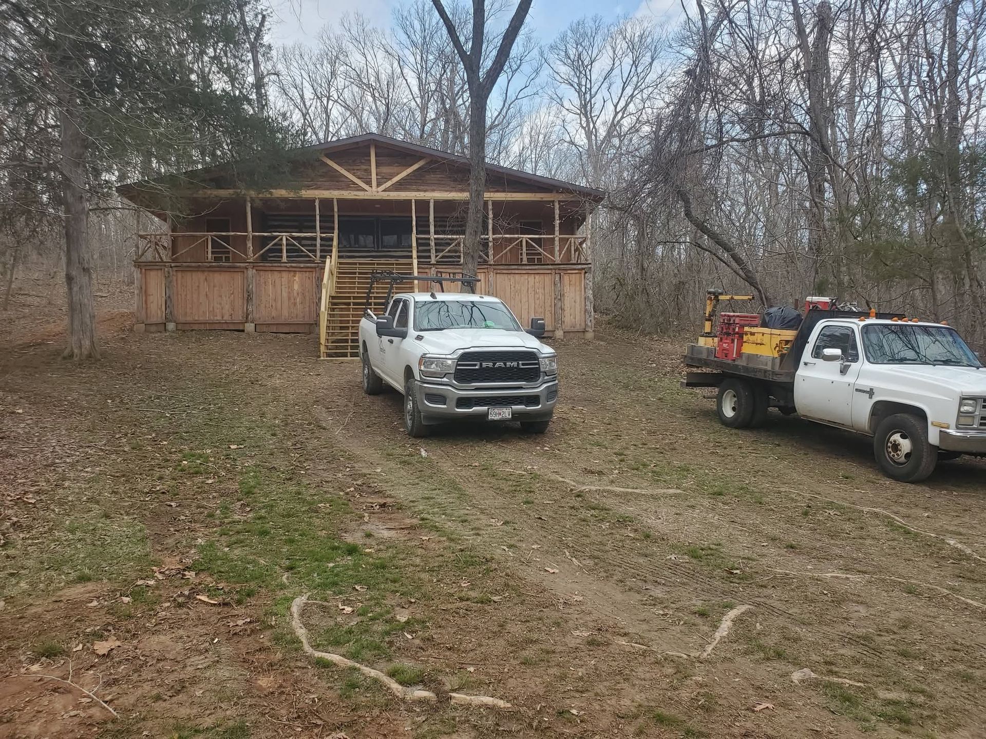 Two pickup trucks parked on a dirt driveway in front of a rustic wooden cabin in the woods