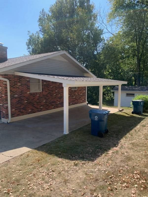 Brick house with white carport, two blue trash bins, and a grassy yard under trees