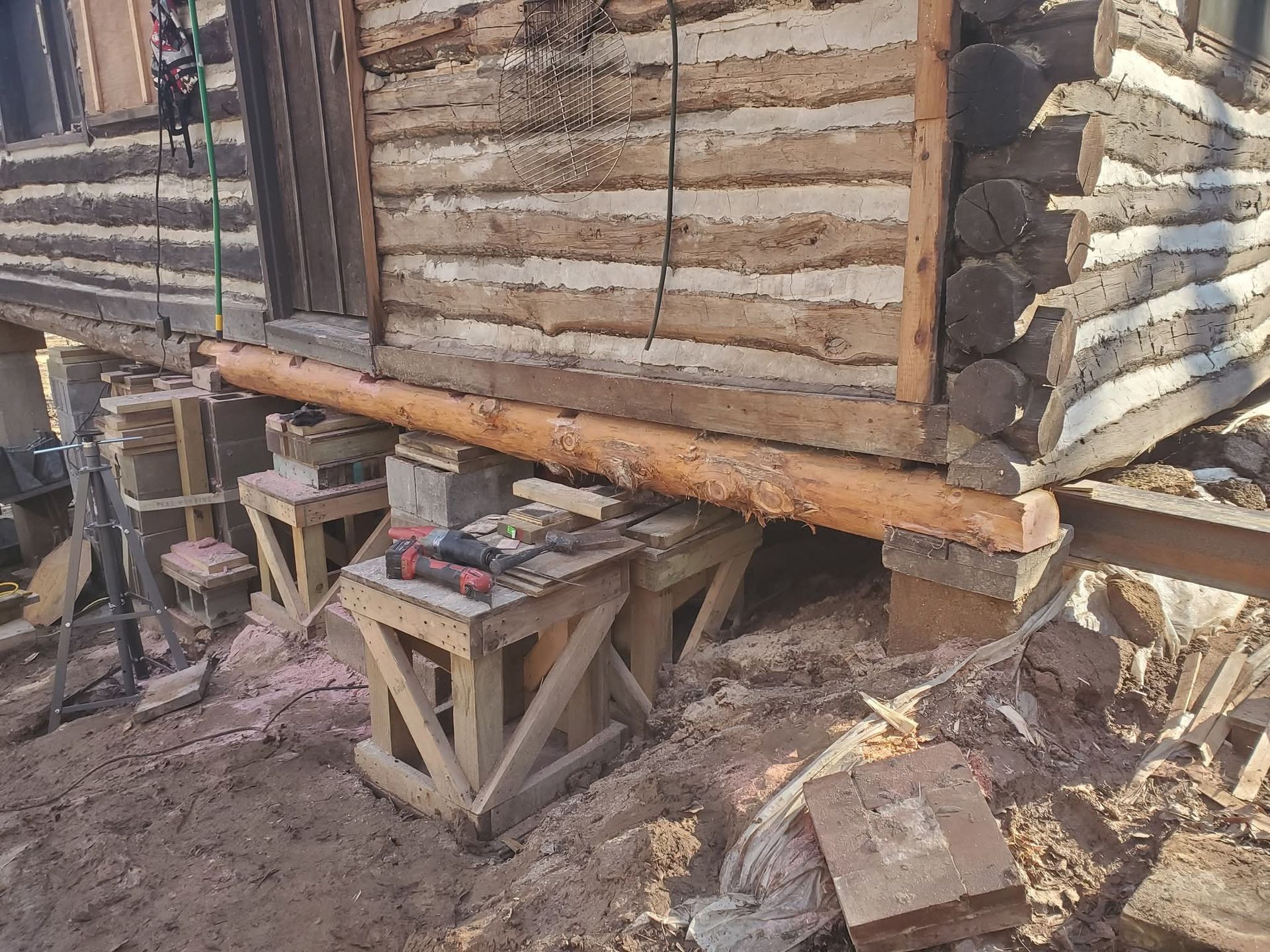 Raised log cabin wall under repair with wooden supports and construction materials on muddy ground