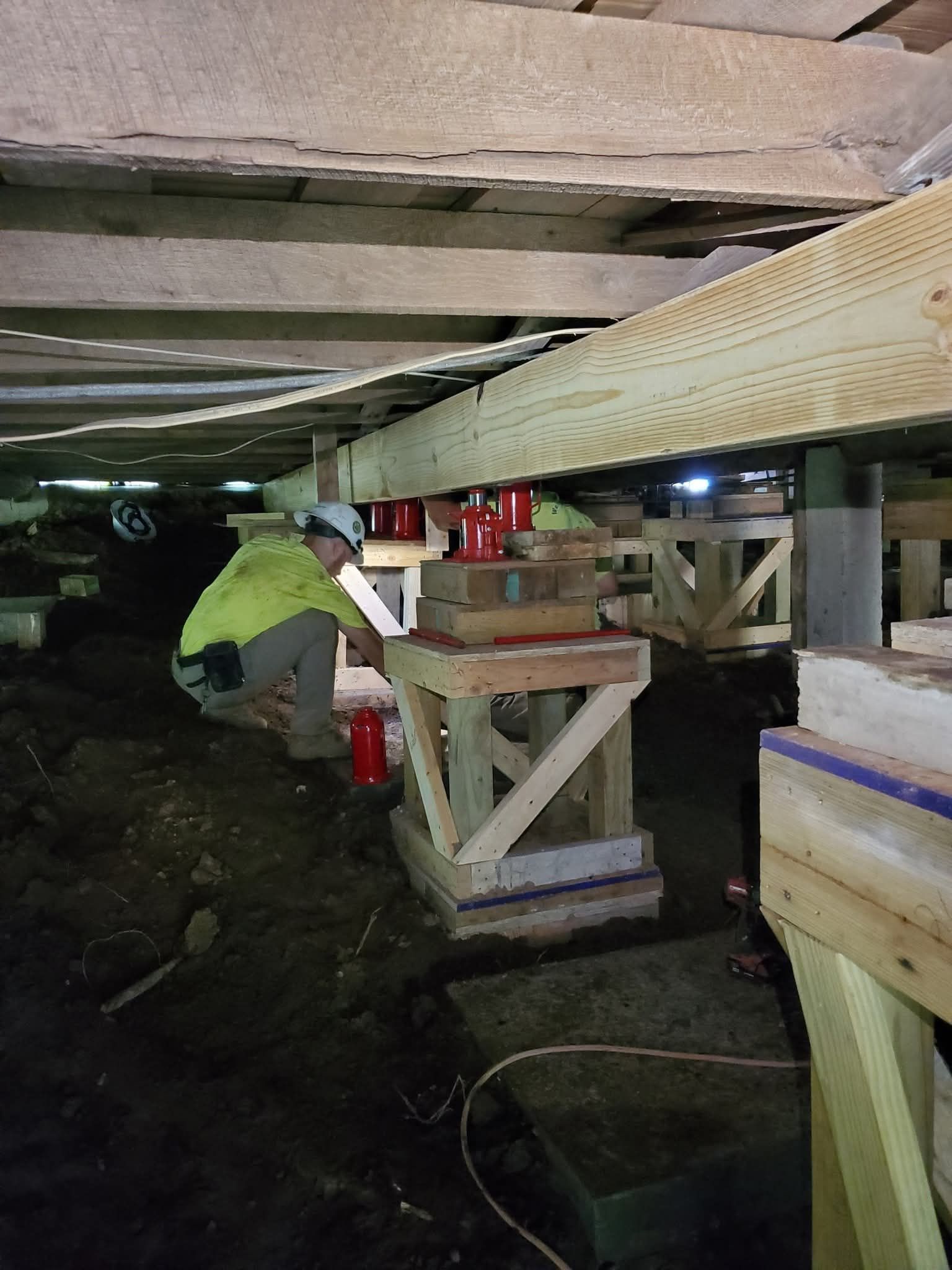 Worker crouching under a house, inspecting wooden beams near tools and supplies.