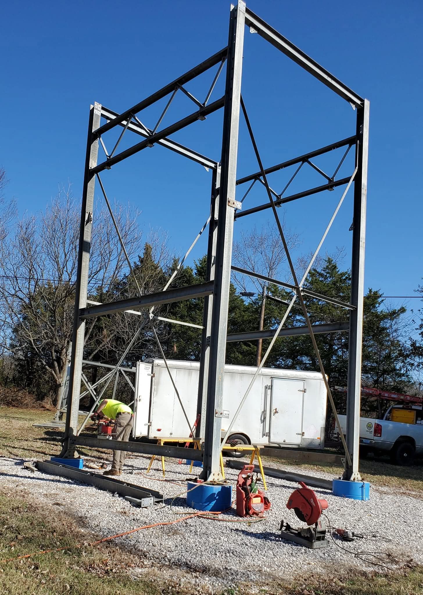 Steel frame under construction on gravel, with workers and equipment beside a white container under a blue sky